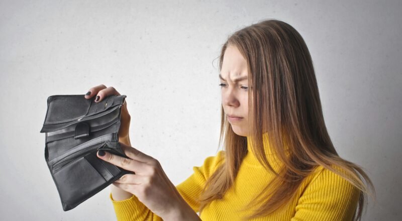 Young woman in a yellow top looking at her empty wallet with a frustrated expression.