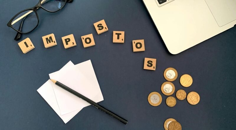 Top view of a workspace with coins, glasses, and the word 'impostos' spelled out, symbolizing taxes.
